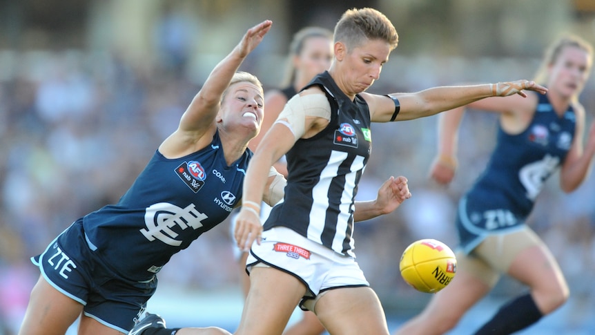 A Collingwood AFLW player tries to get a kick away as a Carlton defender reaches out to tackle her from behind. 