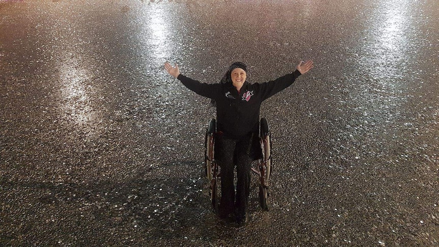 Connie sits in a wheelchair, smiling with arms outstretched, surrounded by hundreds of thousands of five cent coins.