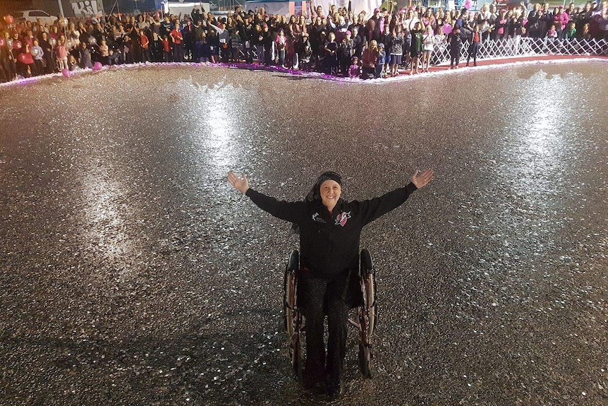 Connie sits in a wheelchair, smiling with arms outstretched, surrounded by hundreds of thousands of five cent coins.