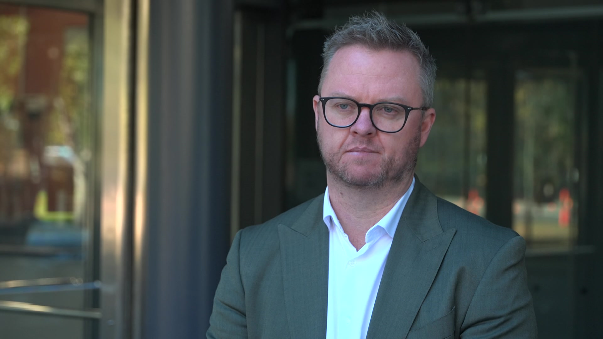 A man in a grey suit and glasses is standing outside a glass building.