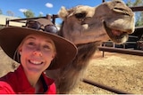 A woman wearing and red shirt and a hat smiling beside a camel