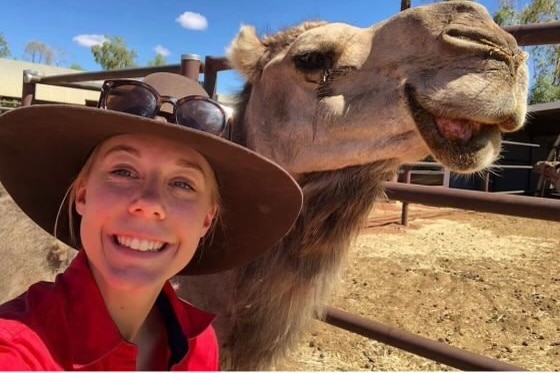 A woman wearing and red shirt and a hat smiling beside a camel