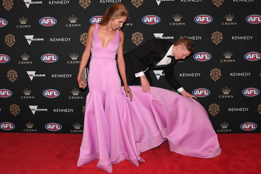Clayton Oliver leans down to lift the trail of his partner's dress on the Brownlow Medal red carpet