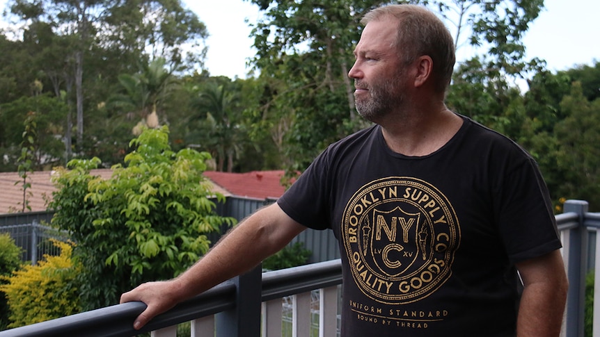 A middle-aged man standing on the back deck of his house, looking out over a green gully behind his property.