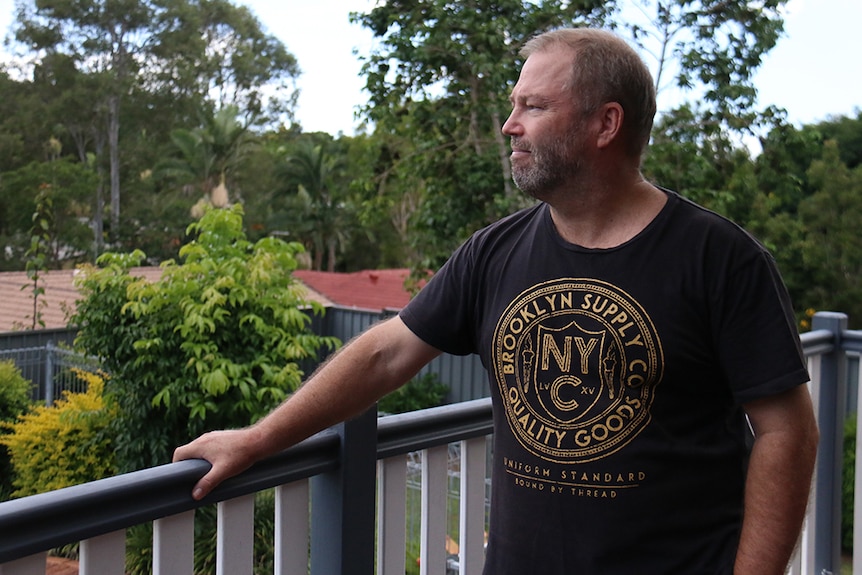 A middle-aged man standing on the back deck of his house, looking out over a green gully behind his property.