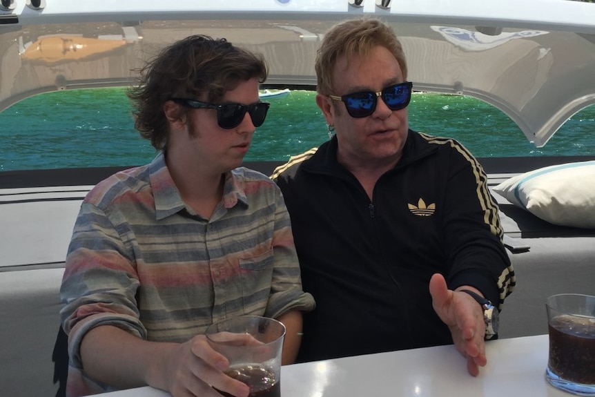 An older man in sunglasses sitting on a yacht and talking with a younger man who is also wearing sunglasses.