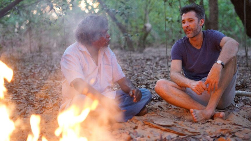 Two men sitting on the ground in the bush chat by a fire