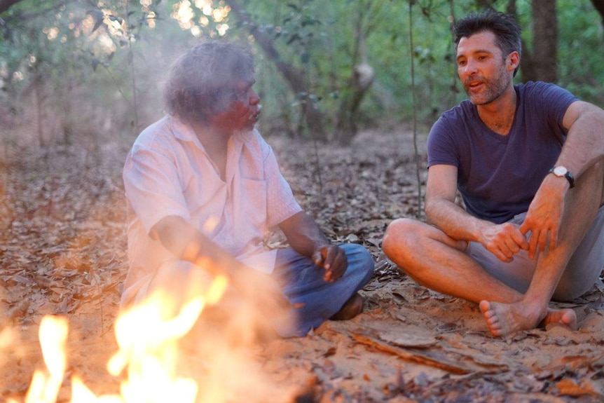 Two men sitting on the ground in the bush chat by a fire