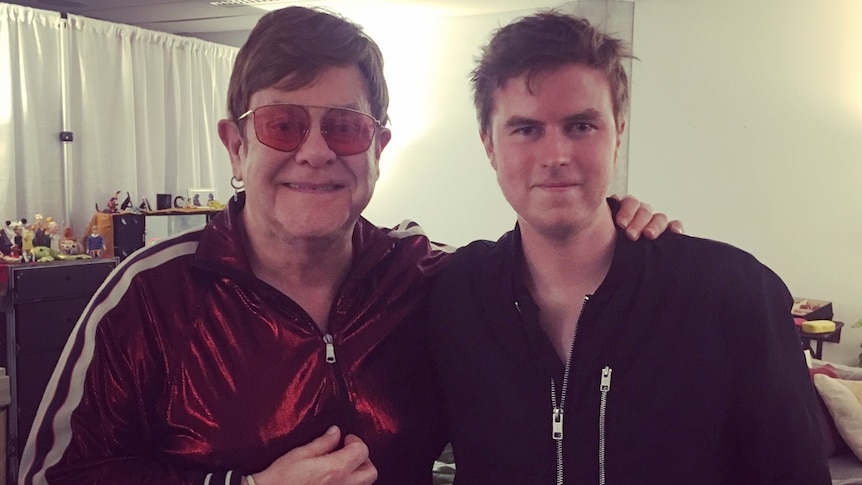 Elton John in a red tracksuit with a younger man in a dark jacket backstage.