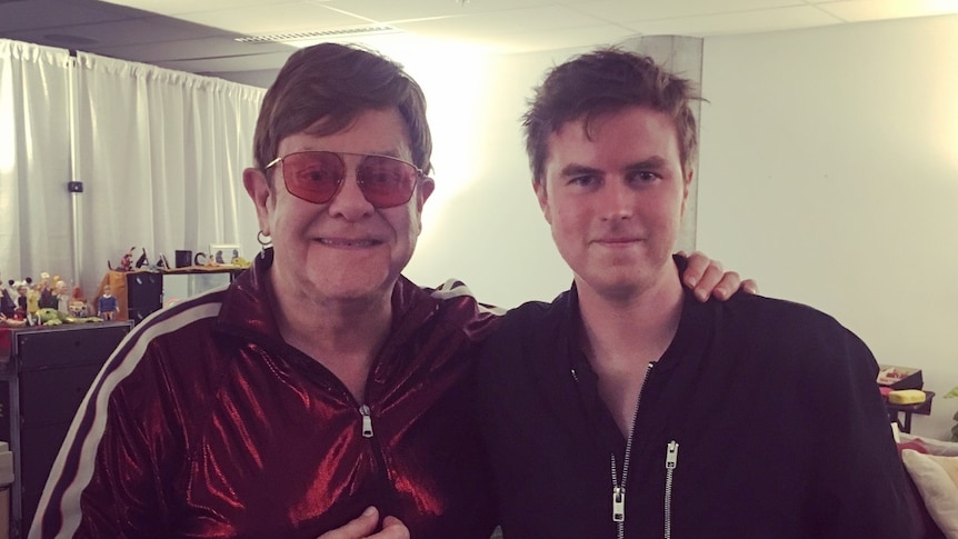 A man in a red tracksuit with a younger man in a dark jacket backstage.