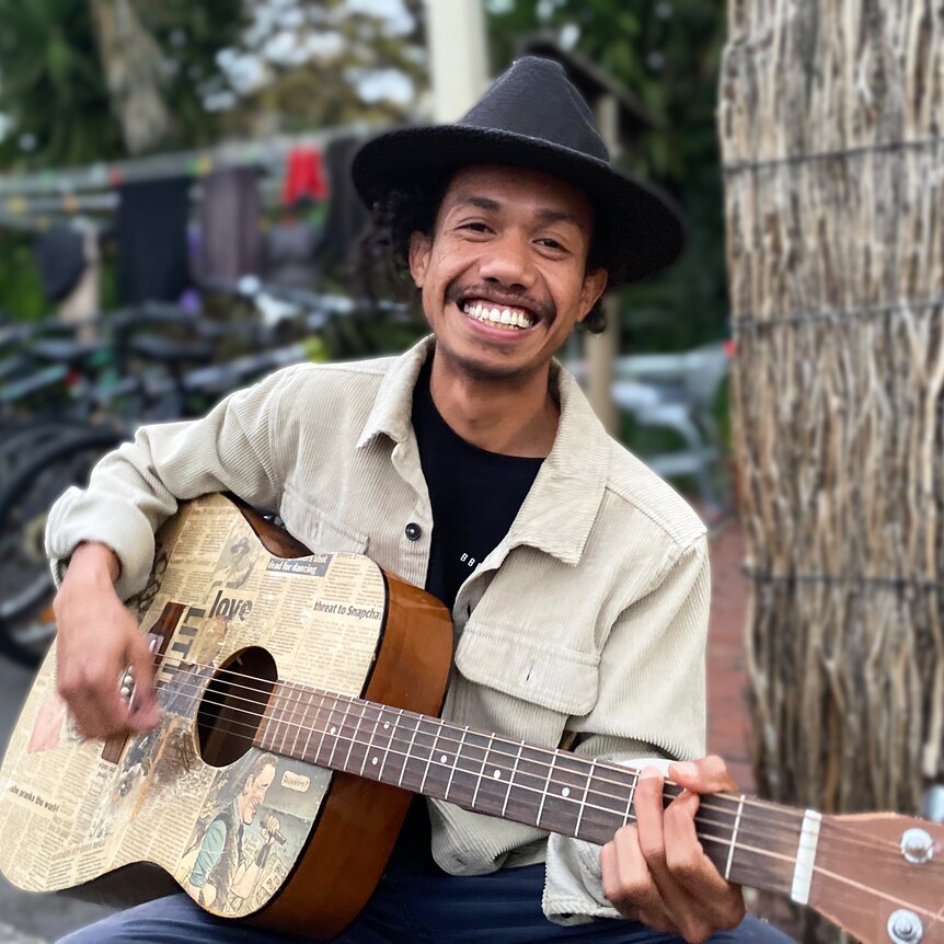 A Timorese man with a cool hat smiles broadly holding a guitar, row of bikes parked behind