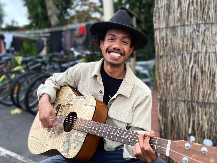 A Timorese man with a cool hat smiles broadly holding a guitar, row of bikes parked behind