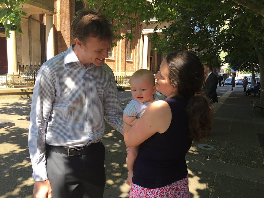 A woman with brown hair holds a baby as her husband looks down at the baby smiling.