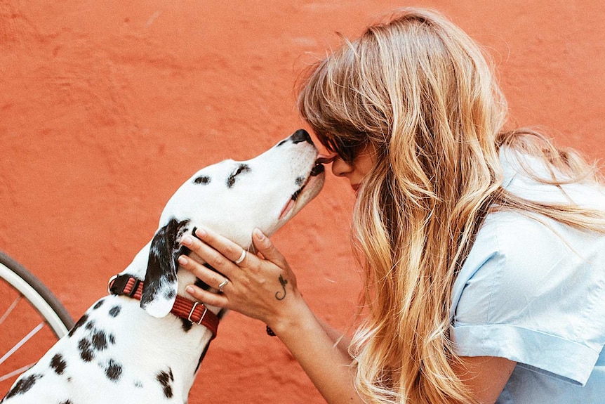 A white dog with black spots licking a blonde woman's face to depict stories of how dogs get people through tough times.