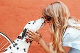 A white dog with black spots licking a blonde woman's face to depict stories of how dogs get people through tough times.
