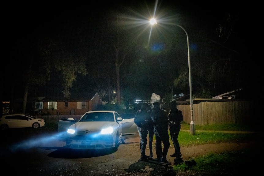 A group of men stand under a street light, one of them is smoking. The bright headlights of a car are shining.