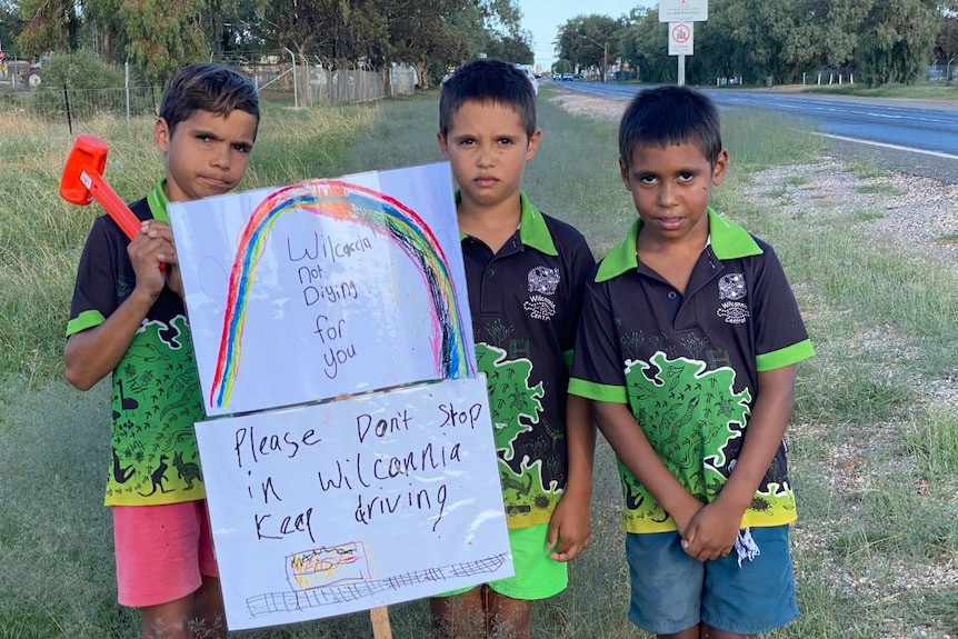 Three boys hold up signs beside a road