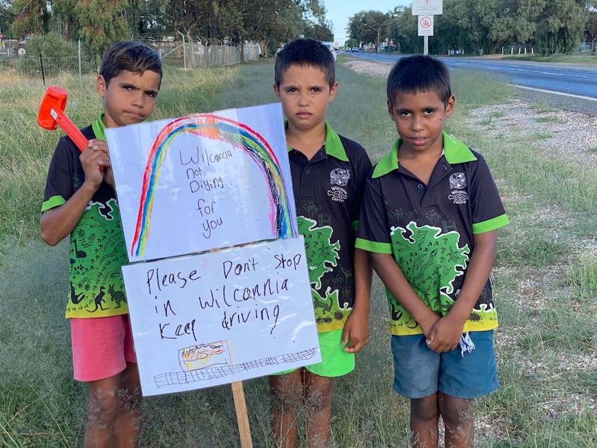 Three boys hold up signs beside a road