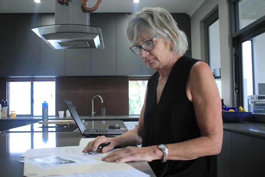 A woman with a black top on and glasses, looks at paperwork on a kitchen bench. She is about 60.