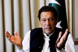 A close up of Imran Khan looking down and gesturing with his hands near a Pakistan flag.