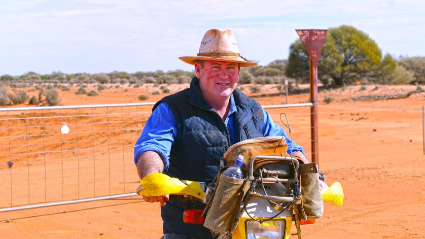 A man wearing an Akubra sits on a motorbike in a paddock