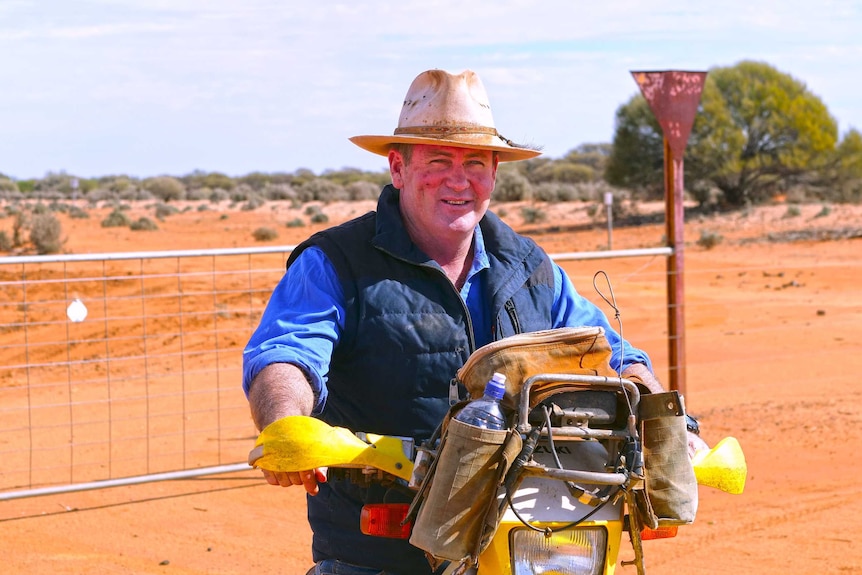 A man wearing an Akubra sits on a motorbike in a paddock