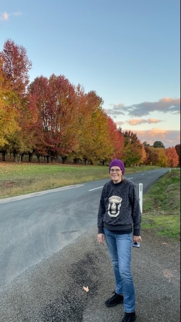 Raylene Herd stands in front of a row of trees as the leaves change in autumn. 