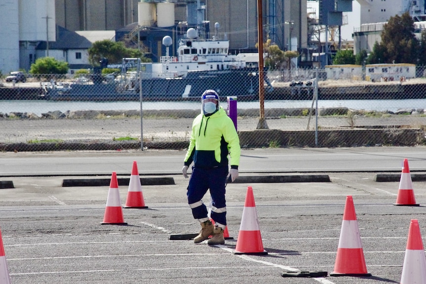 A man wearing a fluro uniform and a face shield surrounded by traffic cones.