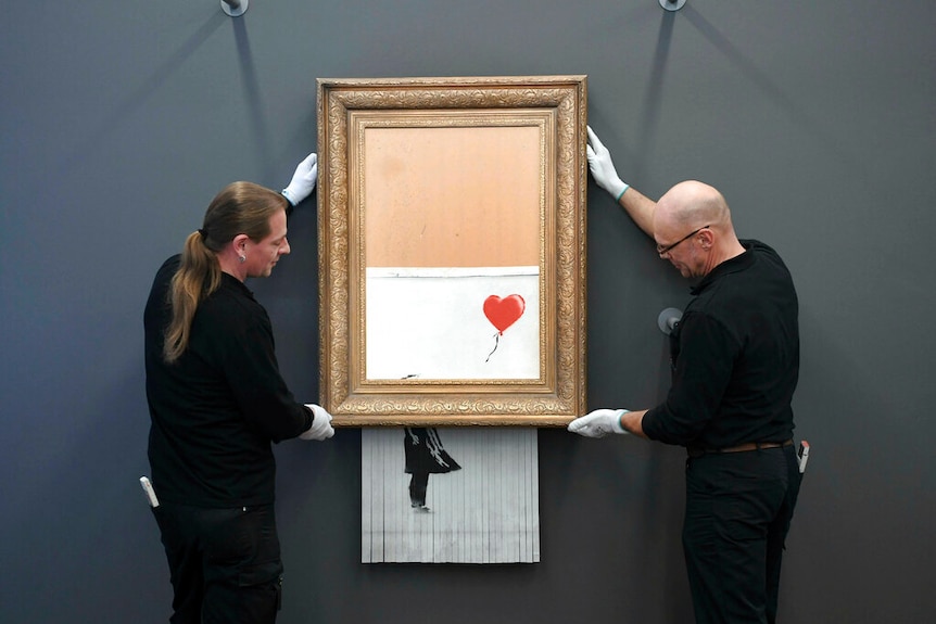 Two men wearing white gloves hang a picture frame with a partially-shredded painting dangling out the bottom.