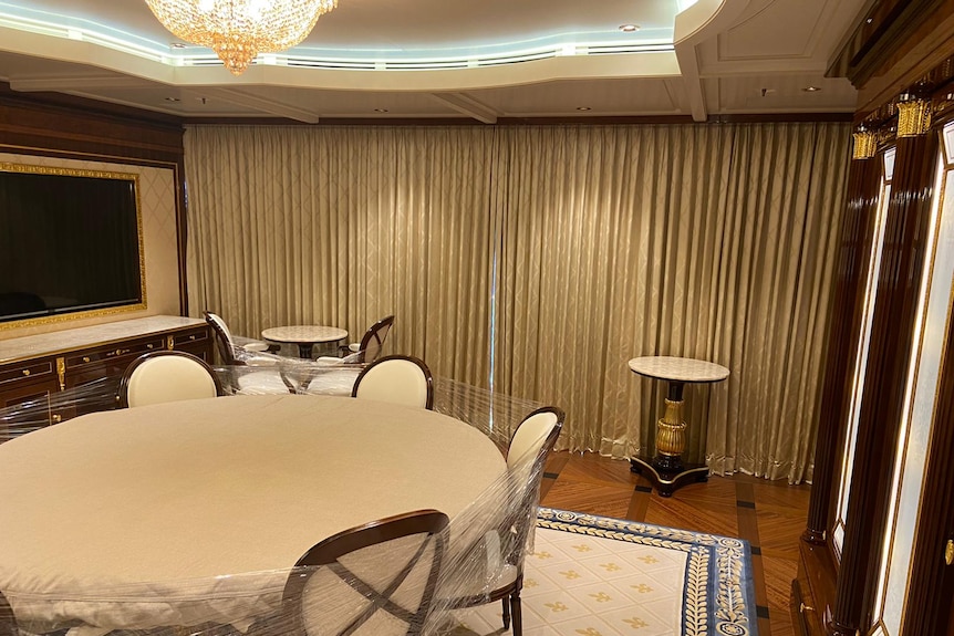 Curtained dining room with cream coloured table and chairs and a large TV on wall and chandelier. 