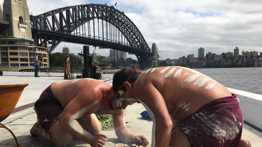 Aboriginal men crouching over smoking embers, with the Harbour Bridge in the background.