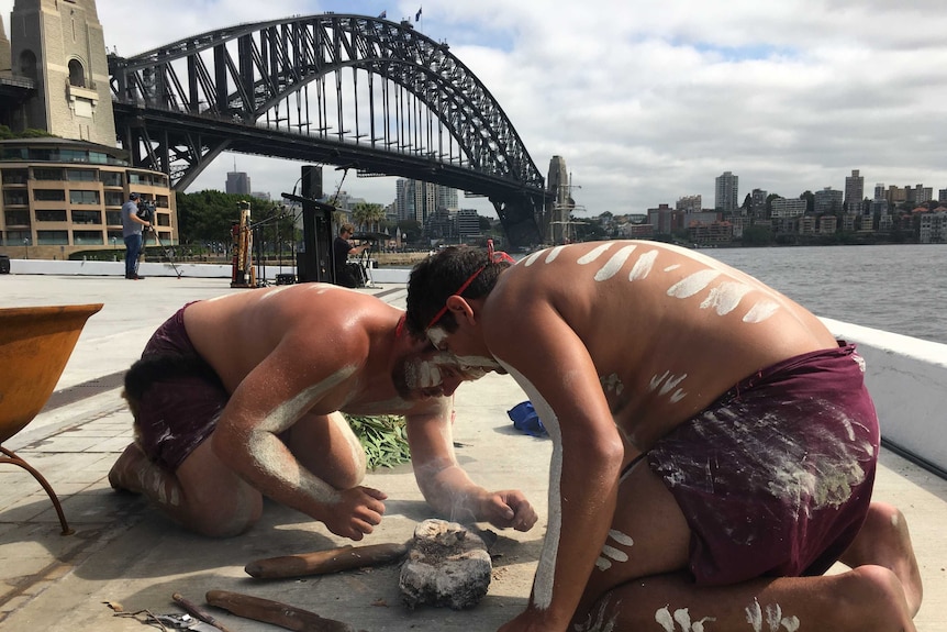 Aboriginal men crouching over smoking embers, with the Harbour Bridge in the background.