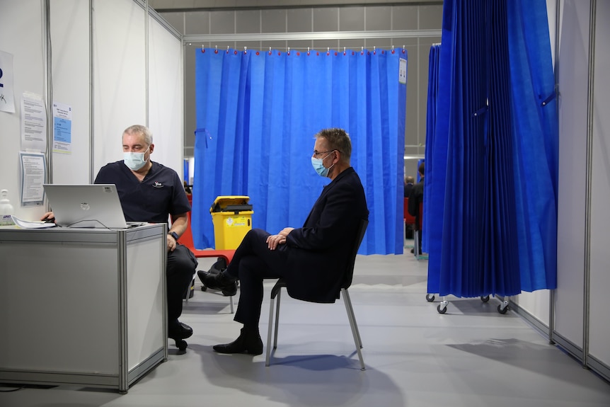 Norman Swan speaks to a vaccine nurse at a vaccination centre with blue curtains in the background.