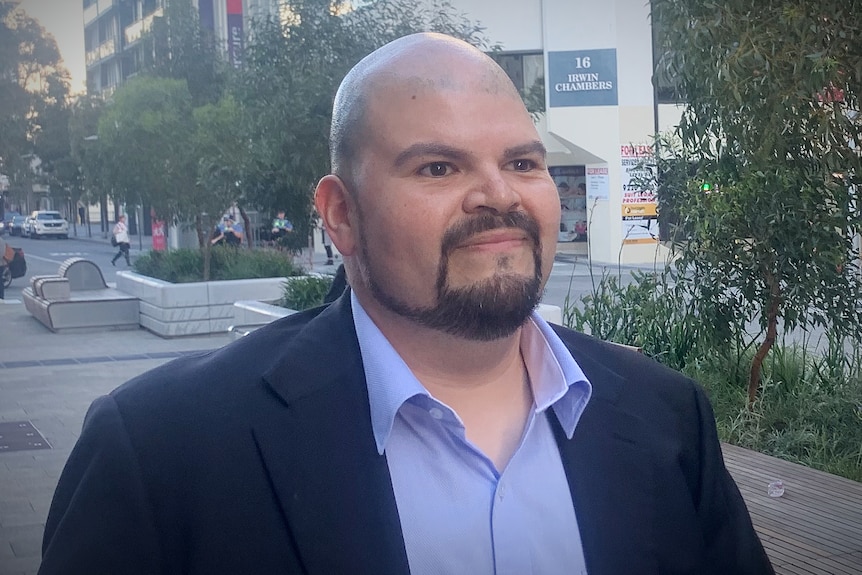 A bald man wearing a suit and blue shirt walks down a city street