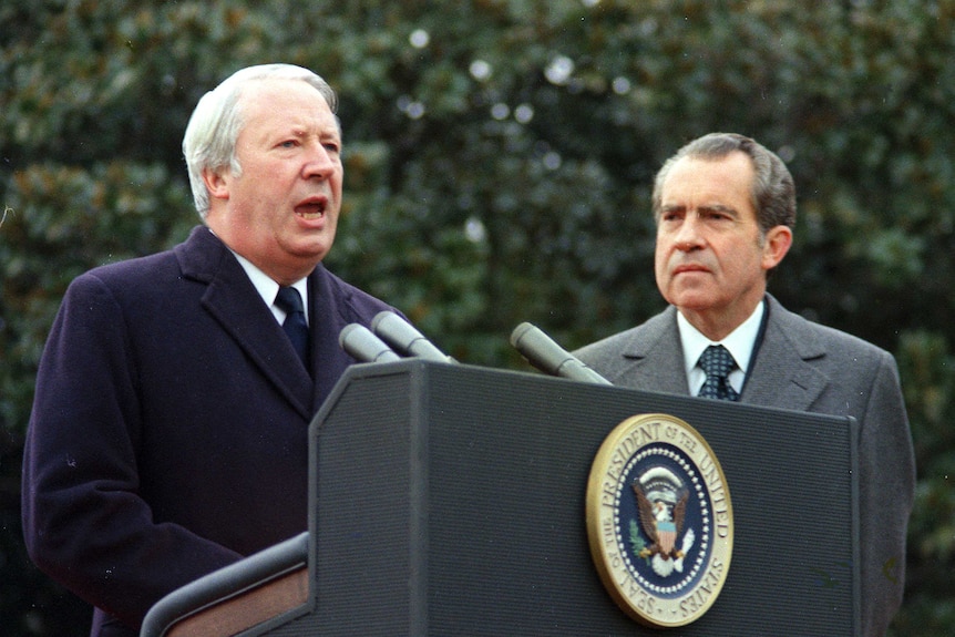 Edward Heath speaks at a podium with the US presidential seal as Richard Nixon looks on.