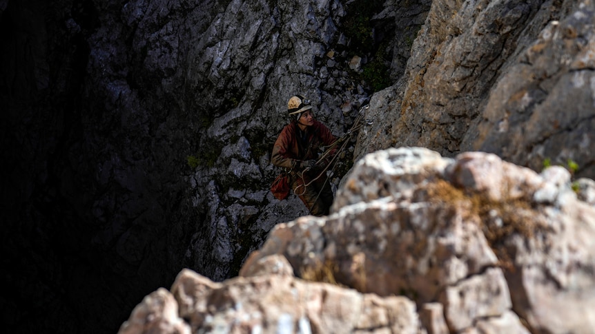 A rescuer wearing a helmet and overalls descends into a cave using a rope