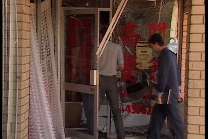 Detectives walking into a restaurant entrance with a shattered window beside them.