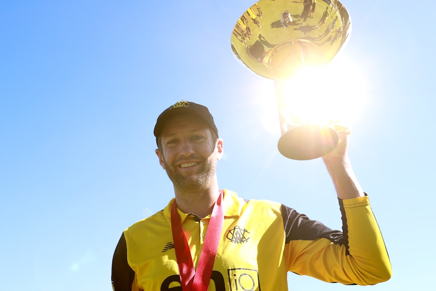 Andrew Tye smiles, with a medal around his neck, holding a gold trophy in the air