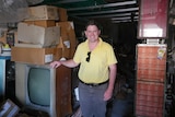 A man in a yellow shirt stands smiling with his hand on an old TV set surrounded by cardboard boxes.