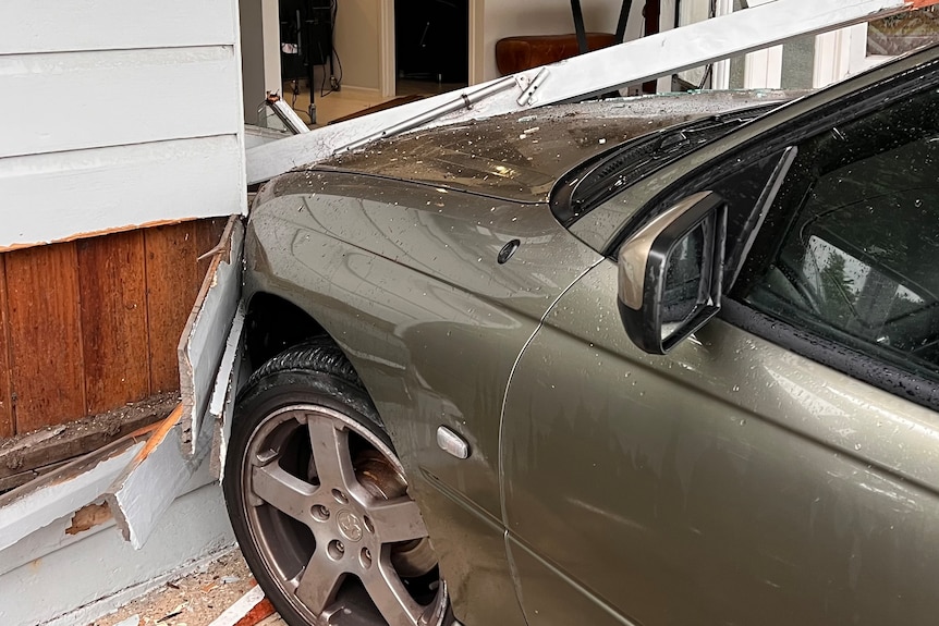 A khaki green metallic car is seen with its bonnet crashed into a shop window, with broken glass around it.