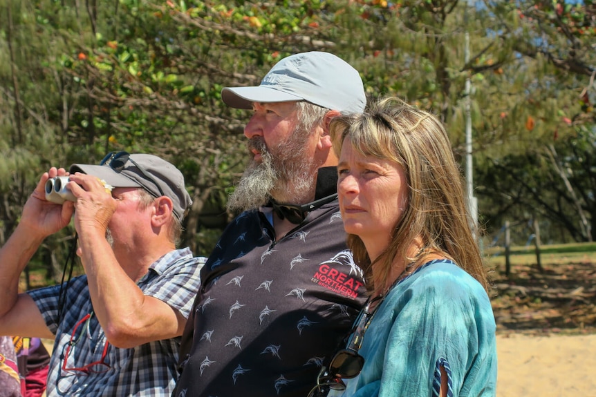 Mark and Kim Oliver look out at the sea where they first spotted an injured whale, a man behind them looks out with binoculars.