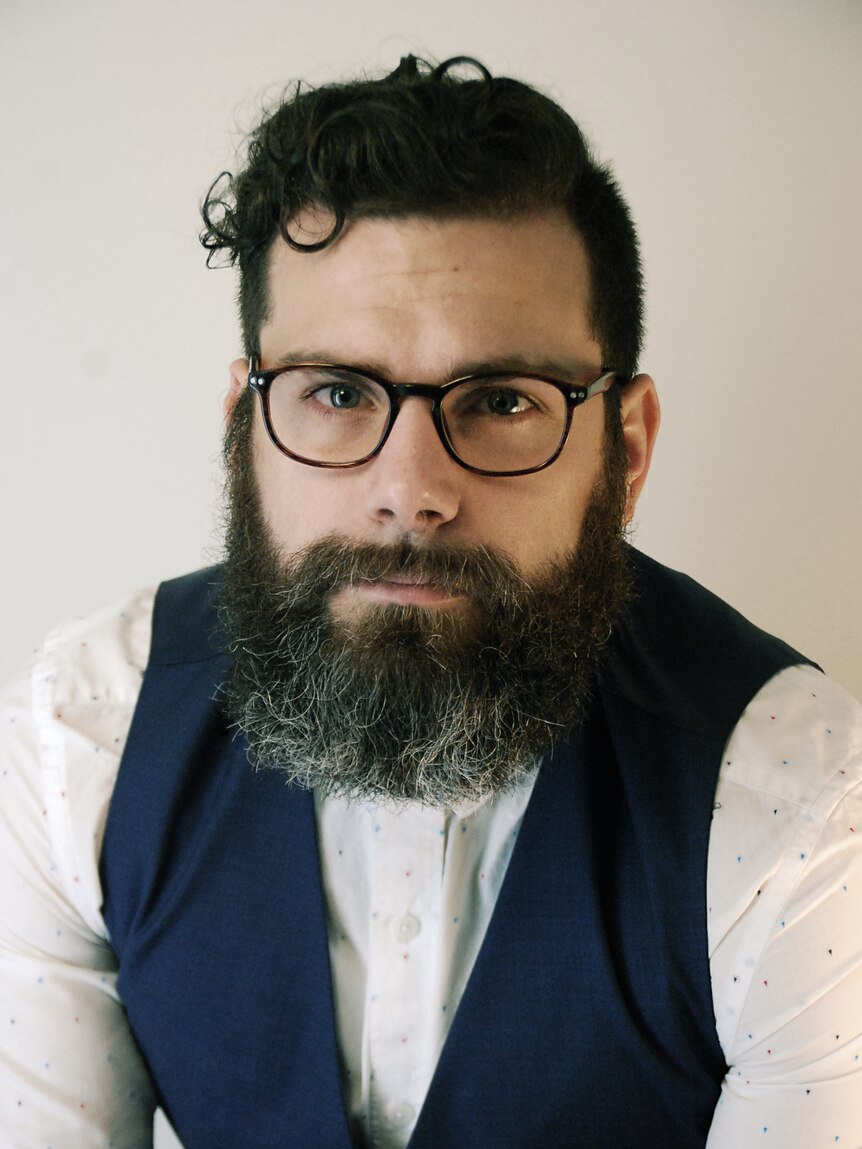 A portrait of a man wearing a white shirt and a blue vest with dark hair, glasses and a beard. 