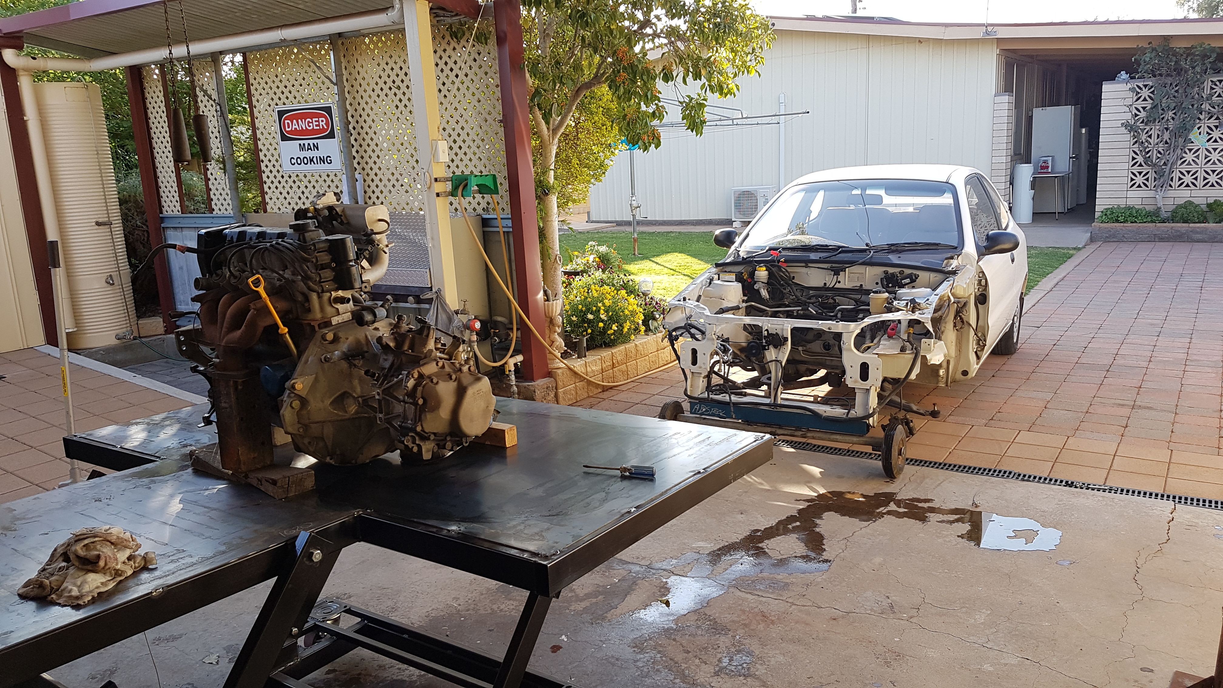 El motor de un coche sobre un banco; al fondo, un coche blanco cuya parte delantera ha sido prácticamente eliminada.