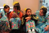 Four girls dressed in blue, floral dance costumes sit on floor. One plays with their phone.