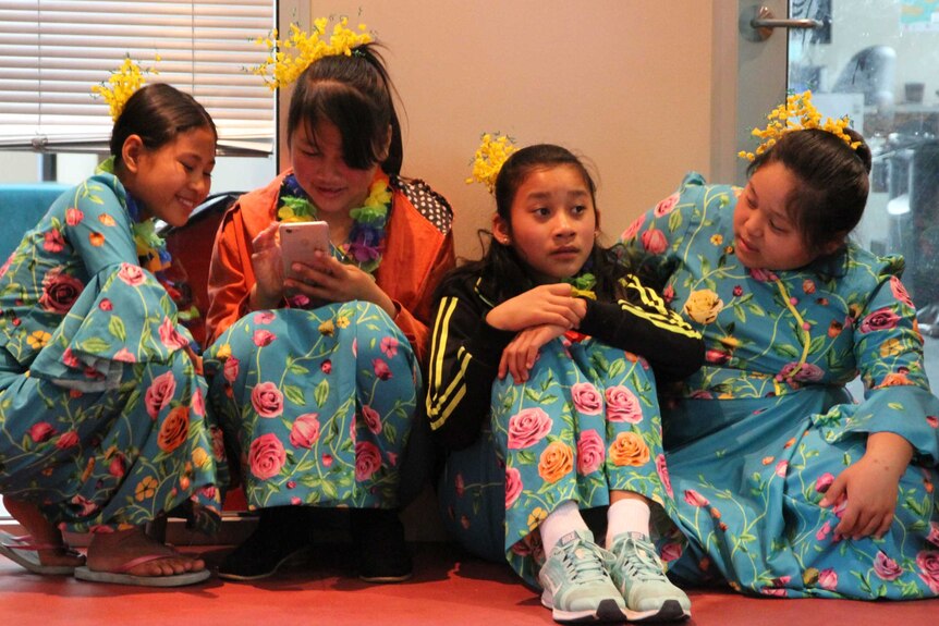 Four girls dressed in blue, floral dance costumes sit on floor. One plays with their phone.