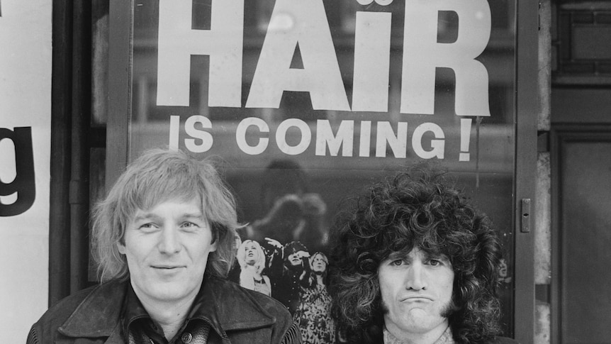 A black and white photo of two men in 1968, the poster in the background says "Hair is coming!"