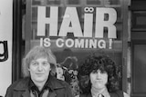 A black and white photo of two men in 1968, the poster in the background says "Hair is coming!"