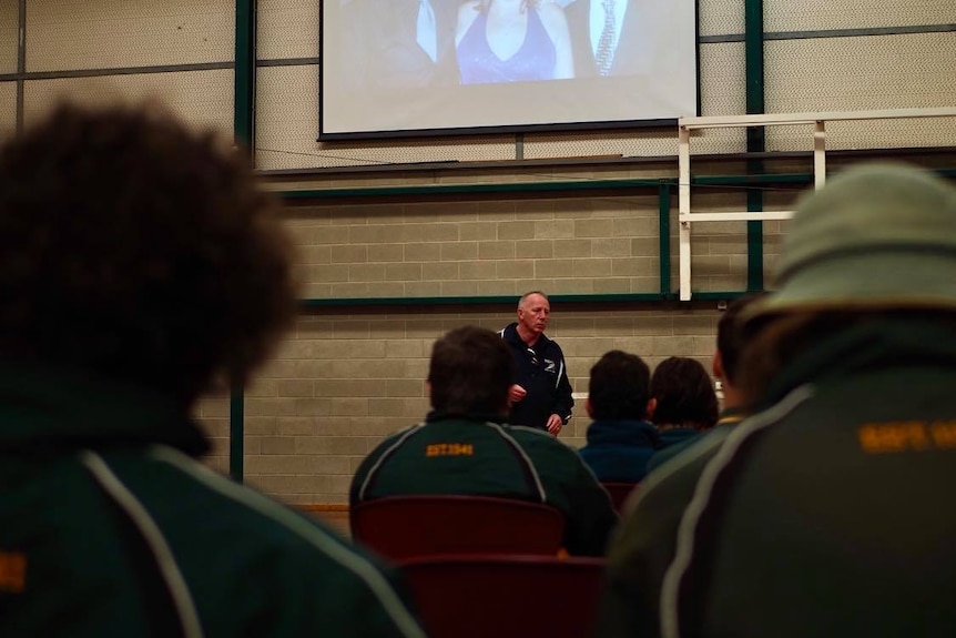 Male school students sit before Neil Davis, who stands in front of the crowd beneath a large projected image of his family.