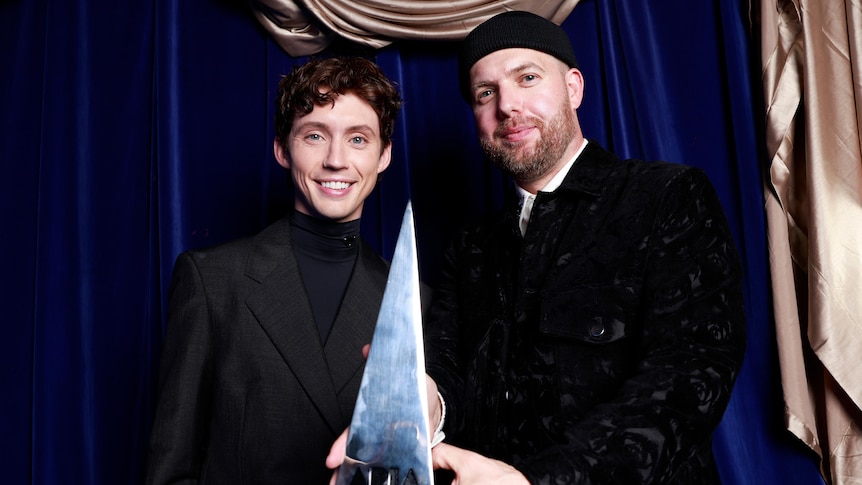 Troye Sivan and Styalz Fuego stand and smile while holding a pyramidal trophy
