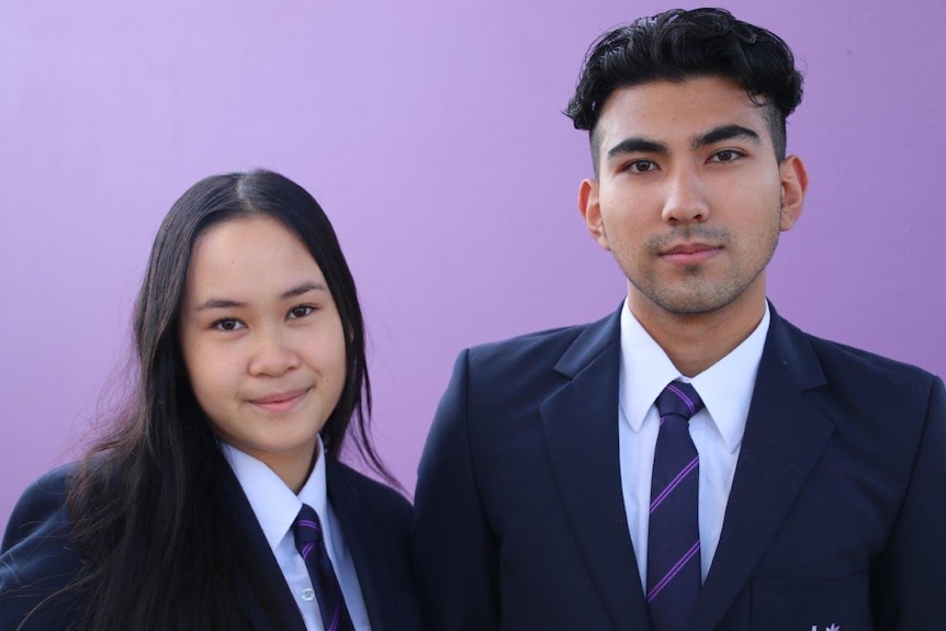 A close up of Kathy and John wearing formal school attire, standing next to each other in front of a purple wall, facing camera